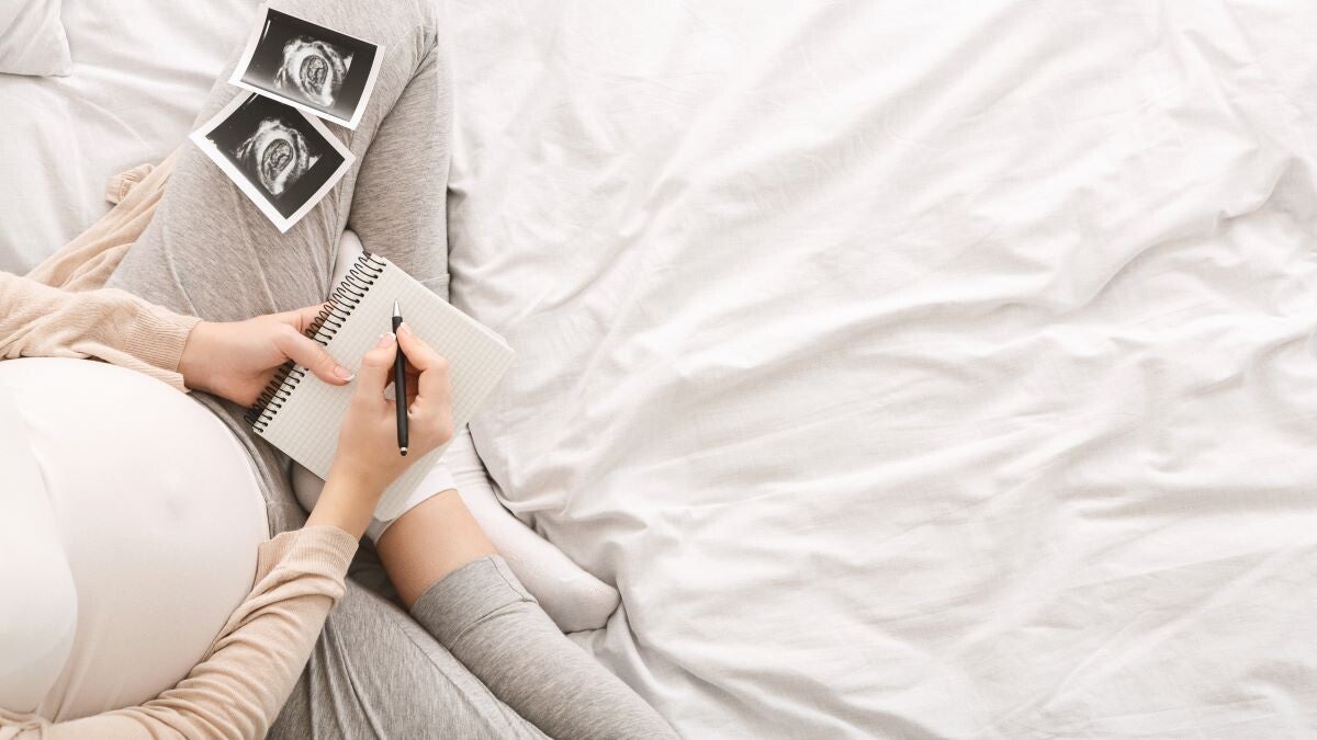 Vogelperspektive einer hochschwangeren Frau, die ein Notizbuch in der Hand hält und zwei Ultraschallbilder ihres Babys