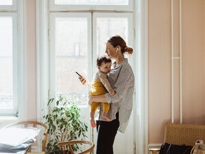 Eine Mutter hält ihr Kleinkind im Arm vor einem sonnenbeschienenen Fenster. Während sie das Kind liebevoll an sich drückt, hält sie ein Smartphone in der Hand.