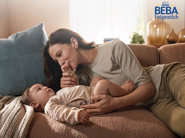 Mutter küsst die Hand des Kleinkinds auf dem Sofa, warmes Sonnenlicht; BEBA-Logo in der Ecke