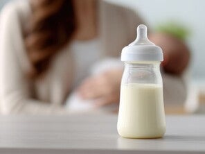 Milchgefülltes Babyfläschchen auf einem Tisch, im unscharfen Hintergrund stillt eine Mutter ihr Baby.