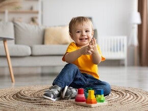 Ein lächelndes Kleinkind sitzt auf einem runden Teppich und klatscht in die Hände vor einem bunten Holzspielzeug.