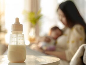 Eine Frau hält ihr Baby und im Vordergrund steht eine Baby Flasche mit Milchinhalt