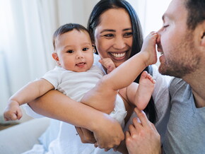 Mutter hält lachend ihr Baby. Das Baby tippt mit dem Fuß die Nase des Vaters an.