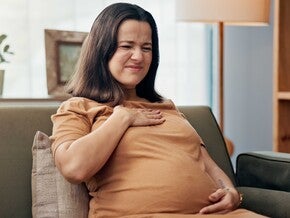 Pregnant woman sitting on a couch, hand on chest, warm light setting.