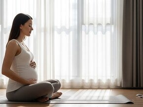 Hochschwangere Frau sitzt im Wohnzimmer auf einer Yoga-Matte und meditiert bei geschlossenen Augen.