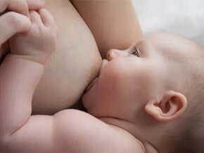 Baby liegt auf dem Arm der Mutter und wird gestillt, während die Mutter die Hand des Babys hält.