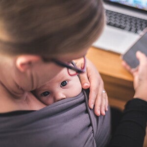 Mutter mit Handy in der Hand und Laptop auf dem Tisch sieht ihr Baby in der Trage an