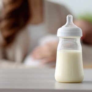 Milchgefülltes Babyfläschchen auf einem Tisch, im unscharfen Hintergrund stillt eine Mutter ihr Baby.
