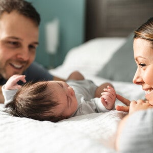 Ein glückliches Elternpaar liegt im Bett und schaut lächelnd ihr neugeborenes Baby an.