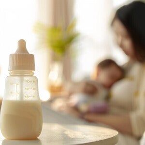 Eine Frau hält ihr Baby und im Vordergrund steht eine Baby Flasche mit Milchinhalt