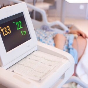Nahaufnahme eines CTG-Monitors, im unscharfen Hintergrund liegt eine schwangere Frau im Bett. 