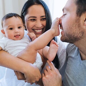 Mutter hält lachend ihr Baby. Das Baby tippt mit dem Fuß die Nase des Vaters an.