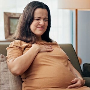 Pregnant woman sitting on a couch, hand on chest, warm light setting.