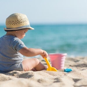Baby trägt zum Schutz vor der Sonne einen Hut, sitzt am Strand und spielt mit Sandspielzeug.