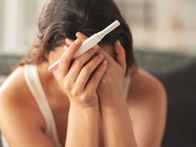 Eine Frau hält einen negativen Schwangerschaftstest und verdeckt aus Verzweiflung ihr Gesicht mit den Händen. 