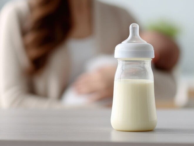 Milchgefülltes Babyfläschchen auf einem Tisch, im unscharfen Hintergrund stillt eine Mutter ihr Baby.