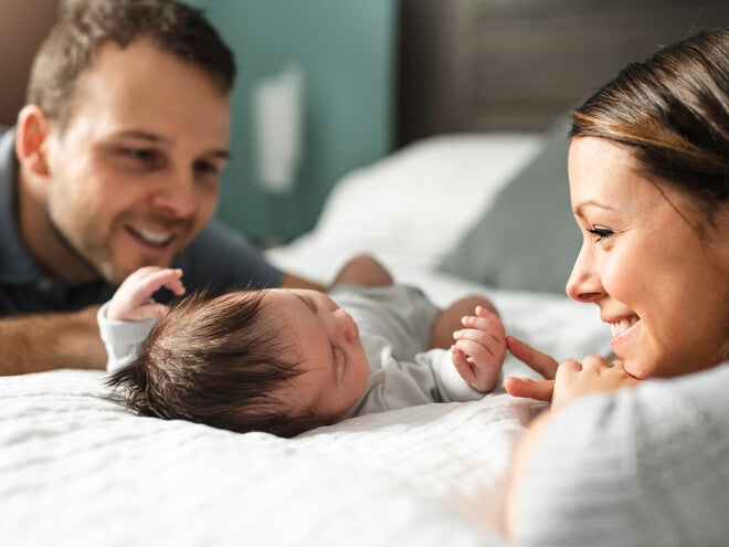 Ein glückliches Elternpaar liegt im Bett und schaut lächelnd ihr neugeborenes Baby an.