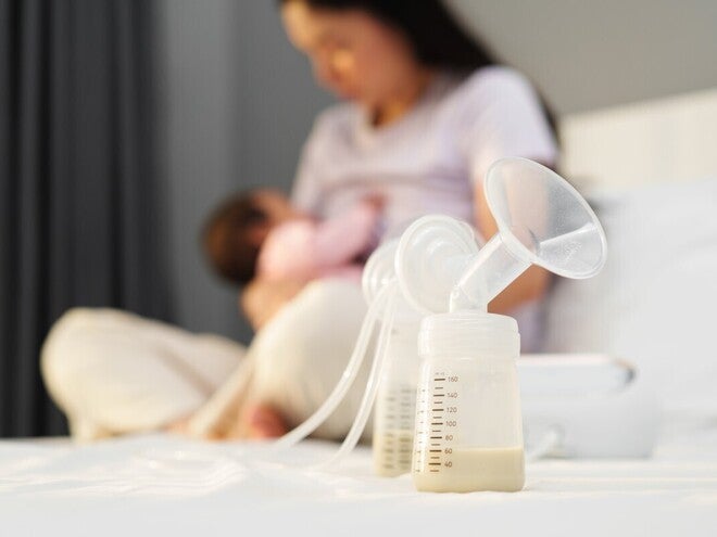 Im Vordergrund steht eine leicht abgefüllte Milchpumpe während die Mutter im Hintergrund ihr Baby auf dem Arm stillt.
