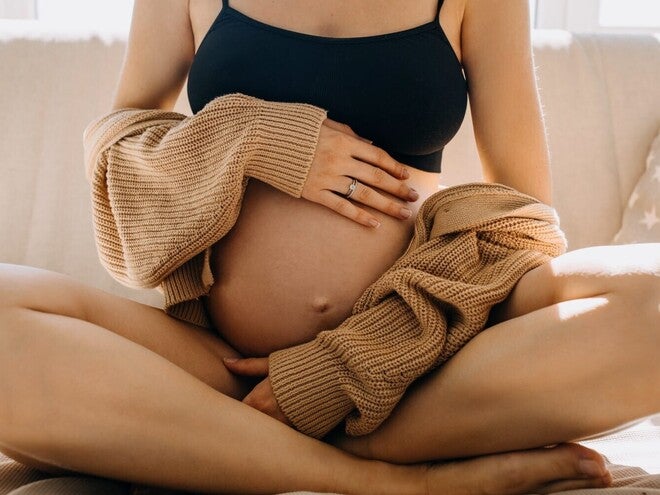 Schwangere Frau sitzt im Schneidersitz auf dem Sofa und berührt sanft ihren Bauch.