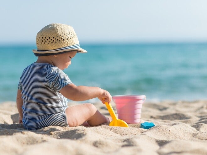 Baby trägt zum Schutz vor der Sonne einen Hut, sitzt am Strand und spielt mit Sandspielzeug.