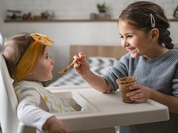 Älteres Kind füttert Baby im Hochstuhl mit Brei – Beikost und Eliminationsdiät bei Babys 