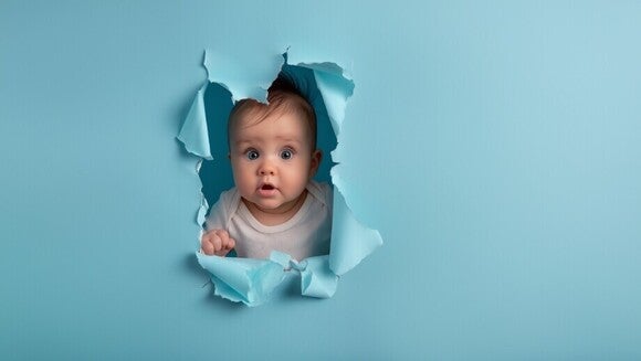 Baby schaut erstaunt durch eine zerrissene, blaue Papierwand durch.