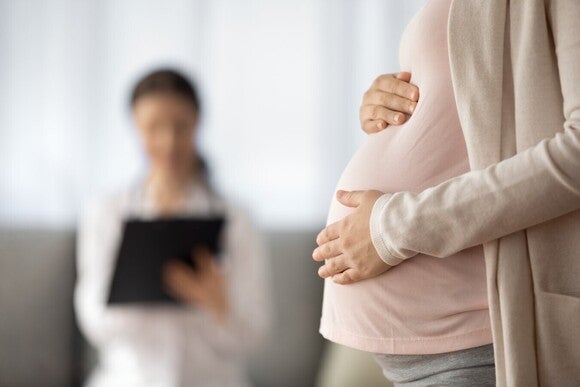 Schwangere Frau hält ihr Hand auf ihren Bauch und im Hintergrund sitzt eine Ärztin