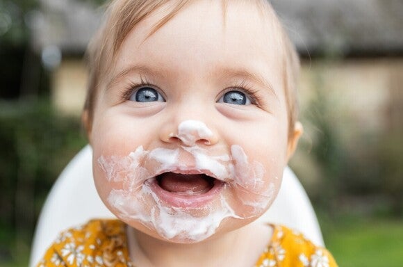 Baby hat Joghurt im Gesicht und strahlt mit großen, blauen Augen und einem Lächeln in die Kamera.