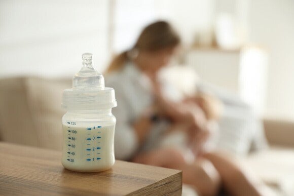Baby Flasche im Vordergrund auf dem Tisch, Mama mit Baby unscharf im Hintergrund zu sehen.