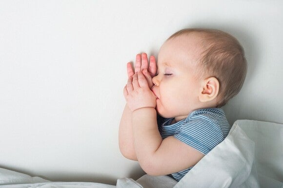 Foto: Schlafendes Baby auf der Seite, Hände am Mund, blauer Strampler, weiße Decke