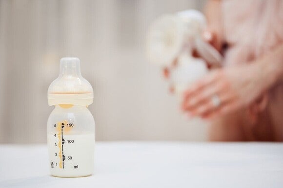 Fläschchen mit Säuglingsmilchnahrung im Vordergrund, Betreuungsperson hält ein Baby im Hintergrund.