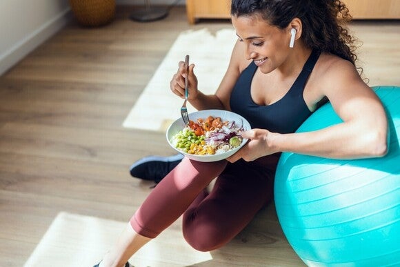 Eine sportlich gekleidete Frau sitzt auf dem Boden, lehnt an einem Gymnastikball und isst eine Salat-Bowl. 