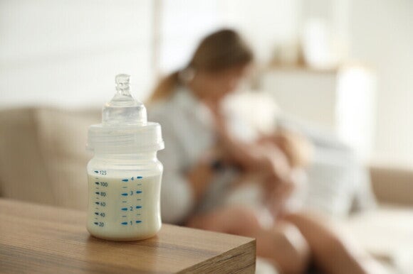 Mutter stillt ihr Baby, während ein Fläschchen mit Muttermilch auf dem Tisch vor ihnen steht Mutter stillt ihr Baby, während ein Fläschchen mit Muttermilch auf dem Tisch vor ihnen steht