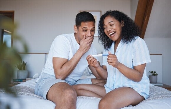 Ein Pärchen sitzt auf dem Bett, sie  sind erfreut und überrascht zugleich, da sie erfahren, dass sie bald ein Baby bekommen.