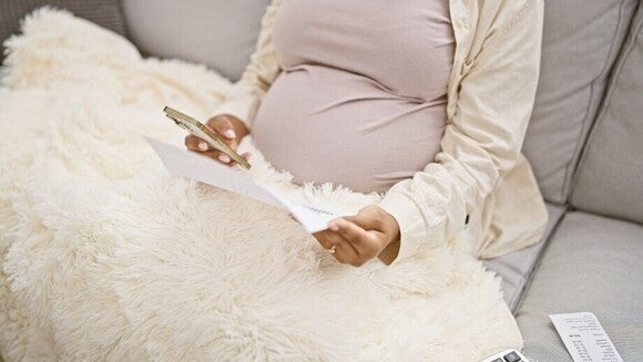 Schwangere Frau sitzt auf dem Sofa mit einer kuscheligen Decke und hält in der einen Hand ihr Smartphone und in der anderen Hand ein Dokument.