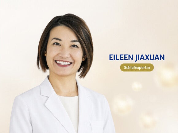 Porträtfoto einer lächelnden Frau in einem weißen Blazer vor hellem Hintergrund, mit blau dargestelltem Namensschriftzug und grünem Abzeichen.