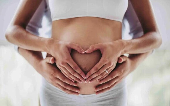 Ein Mann umarmt von hinten eine Schwangere, sie beiden haben die Hände auf ihren Babybauch gelegt