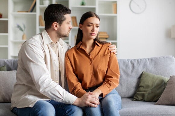 Ein Mann und eine Frau sitzen nach einer Fehlgeburt nachdenklich auf dem Sofa und halten einander