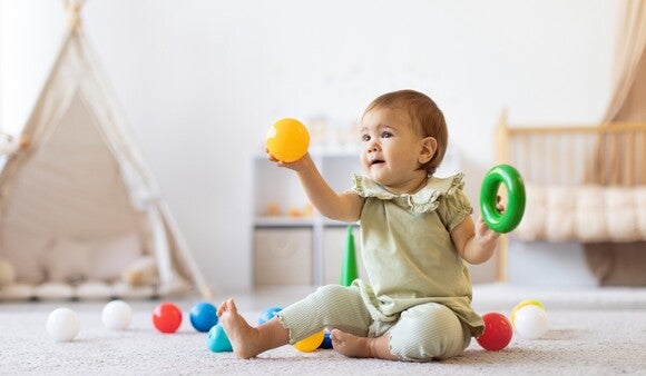 Baby spielt im Kinderzimmer und hält in der rechten Hand einen Plastikball während es in der linken Hand einen Plastik Kringel hält.