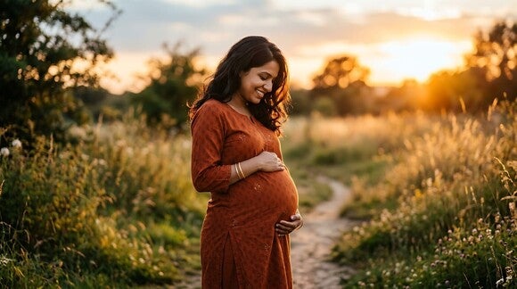 Schwangere Frau hält ihren Bauch und lächelt dabei, während sie bei Sonnenuntergang in der Natur steht.
