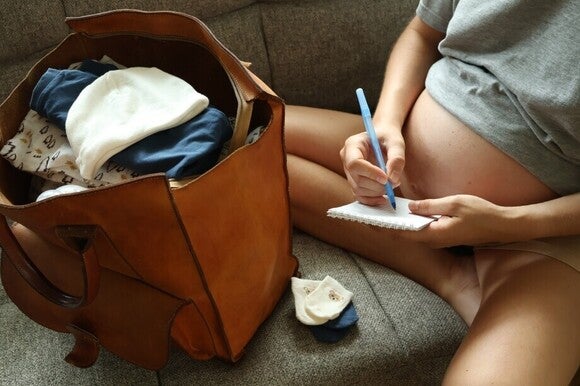Frau sitzt auf der Couch und erstellt neben der gepackten Tasche eine Liste.
