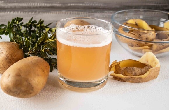 Glass of potato juice with whole potatoes and peels on a table.