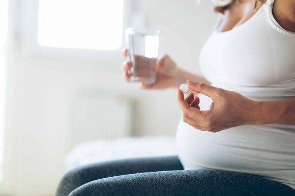 Pregnant woman holding a pill and a glass of water, sitting indoors.