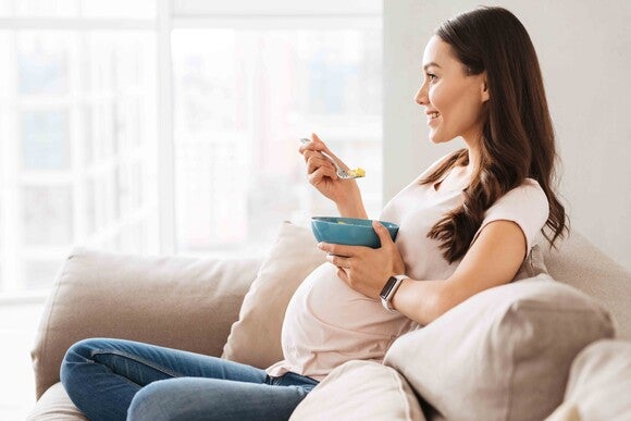 A pregnant woman sitting on a couch, enjoying a bowl of cereal with a relaxed expression.