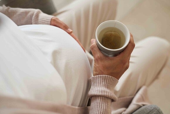Pregnant person holding a cup of tea, sitting indoors in beige clothing.