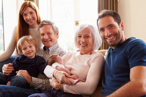 Großeltern, Eltern, Kleinkind und Baby sitzen glücklich zusammen auf einem Sofa - Symbol für die Weitergabe von Genen über Generationen
