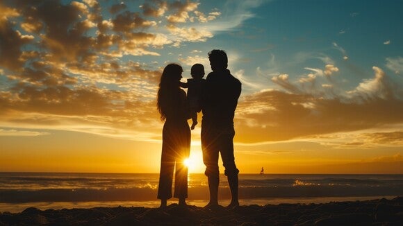 Eltern stehen mit ihrem Baby am Strand vor einem Sonnenuntergang.