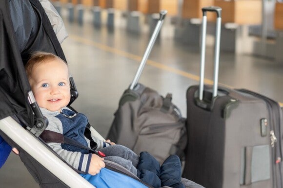 Lächelndes Baby sitzt im Kinderwagen und schaut Richtung Kamera, während daneben zwei gepackte Koffer stehen.
