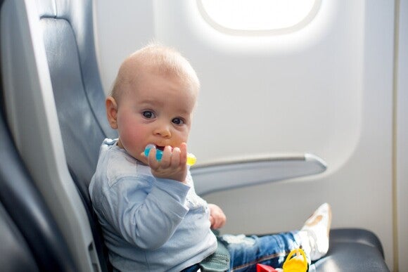 Baby sitzt im Flugzeug angeschnallt auf einem Sitz am Fenster und hält eine Zahnkaustange im Mund fest.