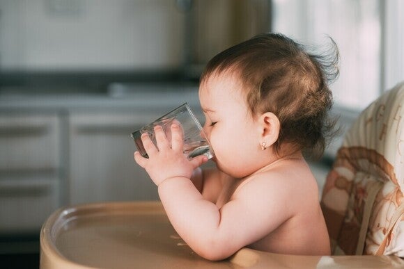 Baby sitzt auf dem Hohlstuhl und trinkt eigenständig Wasser aus einem Glas.